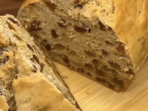 Sliced loaf of spiced fruit bread with visible dried fruit pieces and pale crumb on light wooden surface